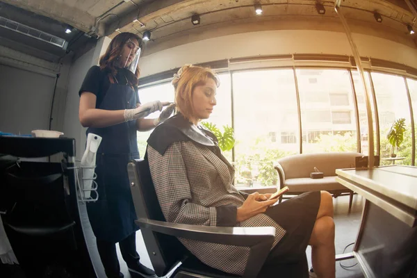 Hairdressers, working in salons with COVID-19 safety measures, are cutting and trimming the hair of a client who wears face shield a salon mask.