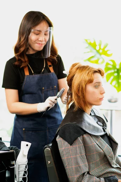 Hairdressers, working in salons with COVID-19 safety measures, are cutting and trimming the hair of a client who wears face shield a salon mask.