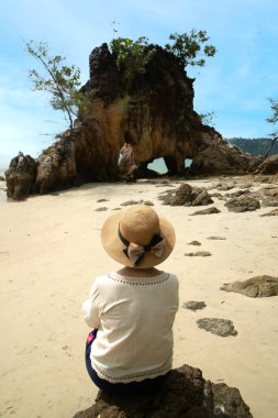 Asyalı kadın turistler sahildeki kayalıklarda oturur ve delikli kayalara ve denize bakarlar. Kumlu sahilde kayalık kayalıklar ve okyanus dalgaları.