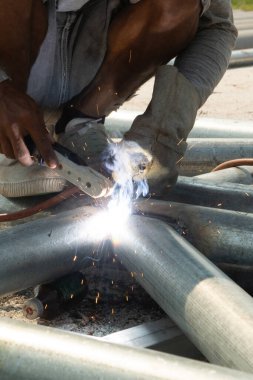 Çelik çatı iskeletinde oturan bir kaynakçı çelik borular ve maske ve deri eldivenler takıyor..