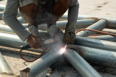 Çelik çatı iskeletinde oturan bir kaynakçı çelik borular ve maske ve deri eldivenler takıyor..