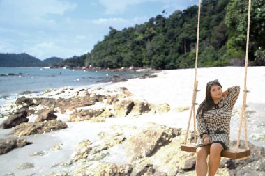 Genç, güzel, Asyalı şişman bir kadın kayalık sahilde salıncakta sallanıyor..