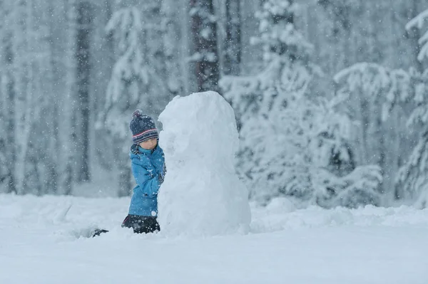 Seorang anak kecil dalam jaket pirus dengan topi biru duduk di lutut patung manusia salju, dengan latar belakang hutan bersalju. konsep tanpa gadget Stok Foto