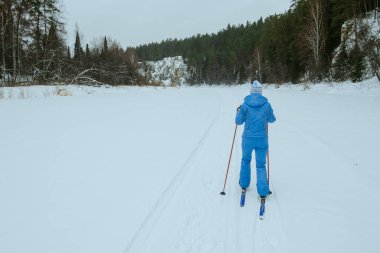 Beyaz şapkalı mavi elbiseli bir kız nehirde kayak yaparken karda yuvarlanıyor.
