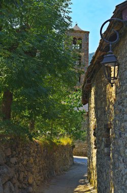 Geleneksel bir Avrupa köyü boyunca uzanan taştan bir sokak yolu, antik duvarları aydınlatan güneş ışığı ve antika bir sokak lambası.