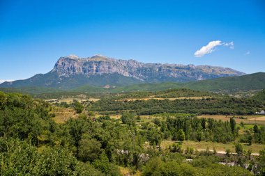 Ainsa vadisi manzarası, açık mavi gökyüzünün altında Pireneler 'in zirveleri olan geniş yeşil tarlaları ve ormanları gösteriyor.