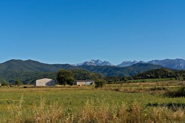 Yeşil tarlalar ve çiftlik binaları açık mavi gökyüzünün altındaki uzak, karlı Pireneler 'e bakıyor.