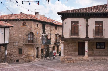 Terracotta çatıları ve ahşap balkonları olan klasik taş binalar renkli sicim ışıklarıyla süslenmiş sakin bir kasaba meydanını çerçeveliyorlar.