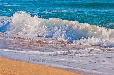Okyanus dalgası kumlu bir sahilde kırılıyor, turkuaz su kıyı şeridinde beyaz köpüğe dönüşüyor. Doğanın gücünü ele geçirmek