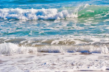 Mavi ve turkuaz okyanus dalgaları ıslak kumda beyaz köpük oluşturur, canlı doğal bir sahneyi betimler.