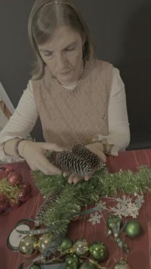 Olgun bir kadın Noel için yeşil bir çelenk üzerinde kozalakları dikkatlice düzenliyor. Tatil işçiliği ve mevsimlik ev dekorasyonu