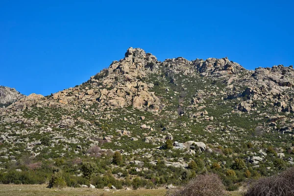 Arid mountain landscape featuring jagged rock formations and scattered green scrub, contrasting with a vibrant clear blue sky