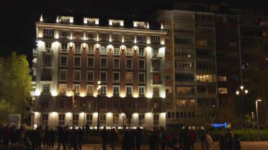 Madrid, İspanya, Noel, 12-14-2025: Kalabalık gece saatlerinde aydınlık bir binanın ön cephesinde kentsel bir parkta yürüyor