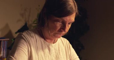 Bir kadın kafası aşağıda bir masada oturuyor. Beyaz bir gömlek giyiyor ve boynunda bir kolye var.