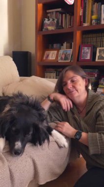 Bir kadın kanepede siyah beyaz bir köpeği okşuyor. Köpek kanepede uzanıyor ve kadın yerde oturuyor. Sahne rahat ve rahatlatıcı.