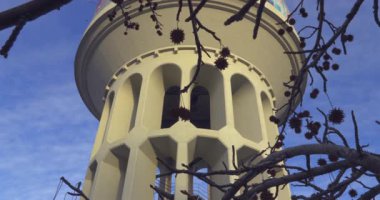 Tepesinde çan olan uzun beyaz bir kule. Kule ağaçlarla çevrili ve gökyüzü mavi.