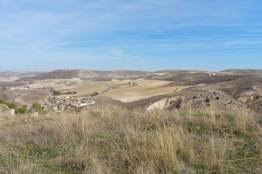 Uzaklarda birkaç evi olan geniş, açık bir arazi. Gökyüzü açık ve mavi
