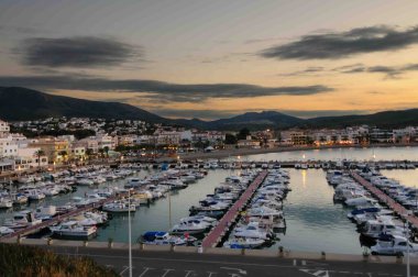 Birçok teknenin kenetlendiği güzel bir marina manzarası. Gökyüzü bulutlu ve güneş batıyor.