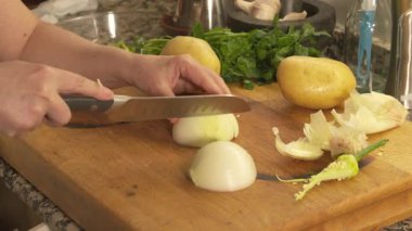 Tahta kesme tahtasıyla taze beyaz soğan kesen kadın elleri. Yanında patates, maydanoz ve sarımsak gibi malzemeler var.