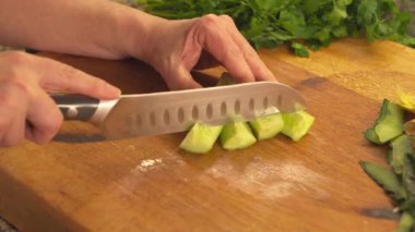 Female hands holding a sharp kitchen knife and chopping a green cucumber into small pieces. A close up view of meal preparation on a board