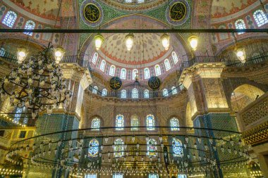 Görüntü, karmaşık mimari ayrıntılar, renkli vitray pencereler ve dekoratif avizelerle karakterize edilen bir caminin içini gösteriyor. Tavan dekoratif desenlerle kaplı..