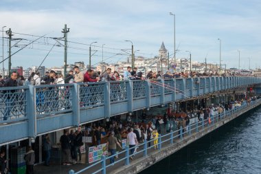 İstanbul 'da kalabalık bir köprüde balık tutan insanlar yer alıyor. Satıcılar liman bölgesi boyunca mevcut, binalar ve arka planda açık bir gökyüzünün altında görünen bir kule var..