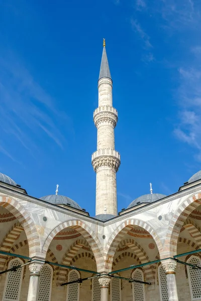 Yüksek bir cami minaresi, açık mavi gökyüzüne karşı, kubbeleri olan kemerli bir yapının üzerinde yükselir. Yüzeyde dekoratif unsurlarla mimari ayrıntılar görünür..