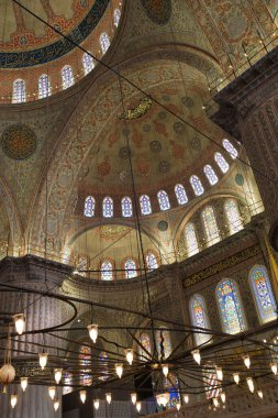 Resimde, tarihi bir caminin karmaşık tavanı ve boyalı camları, duvarlarda avizeler ve dekoratif desenler yer alıyor..