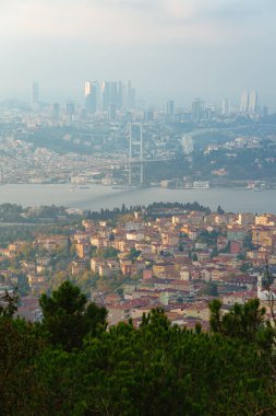 İstanbul 'a ait yerleşim binaları ve su kütlesi genişliğinde bir köprünün yer aldığı hava perspektifi. Şehir kısmen sisle kaplı ve arka planda modern gökdelenler görülüyor..