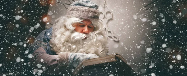 青いカフタンの魔法のロシアのサンタクロース 祖父フロスト はクリスマスツリーの前に座って 大きな開きの願い事の本を読んでいます パノラマのバナー クリスマスのコンセプト ストックフォト