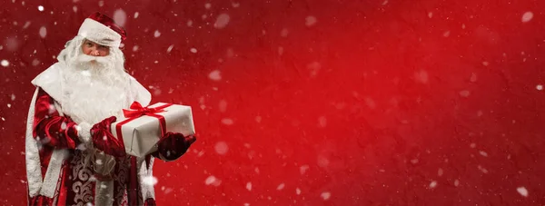 お祝いの赤い新年の背景に雪の結晶と赤いリボンで結ばれた贈り物の白い箱を持つ赤いカフタンのロシアのサンタクロース Ded Moroz コピースペース付きのパノラマバナー ストック写真