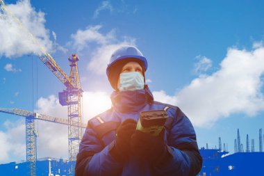 Male engineer surveyor in blue helmet and winter jacket stands in front of building under construction in protective medical mask from coronavirus with a tablet from GPS. Cold clear day. Mixed media clipart
