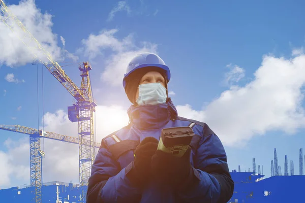 青いヘルメットと冬のジャケットの男性エンジニア測量士は Gpsからタブレットでコロナウイルスから保護医療マスクの建設中の建物の前に立っています 寒い晴れた日 混合メディア ロイヤリティフリーのストック写真