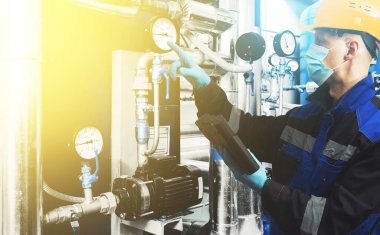 Korunaklı miğfer, maske ve eldiven takan erkek mühendis Coronavirus 'a bağlı, dokunmatik kontrol cihazı endüstriyel kazan dairesini kontrol ediyor. Borular, boru hatları, manometreler, gaz dağıtım ünitesi. Karışık ortam