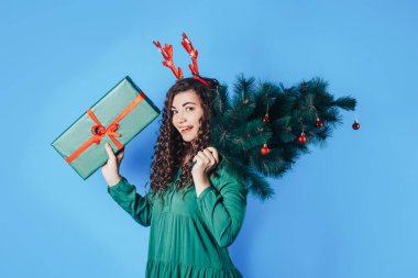 Elinde büyük bir hediye ve elinde bir Noel ağacı tutan genç bir kadın. Mavi arka planda boynuzları var..