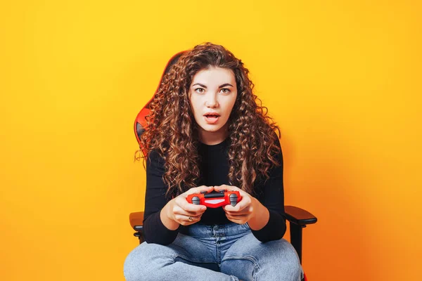 Woman sitting behind gaming chair in her hands holding red gamepad on yellow background.