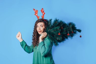 Elinde Noel ağacı tutan genç bir kadının mavi arka planda kırmızı geyik boynuzları var..