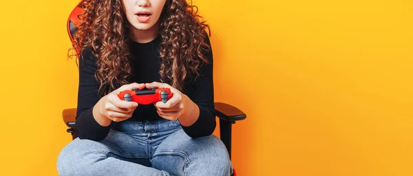 Woman sitting behind gaming chair in her hands holding red gamepad on yellow background.