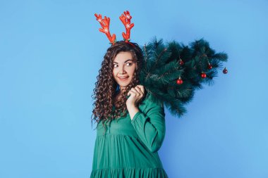 Elinde Noel ağacı tutan genç bir kadının mavi arka planda kırmızı geyik boynuzları var..