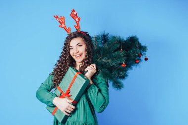 Elinde Noel ağacı ve kutu tutan genç bir kadın. Mavi arka planda kırmızı geyik boynuzları var..