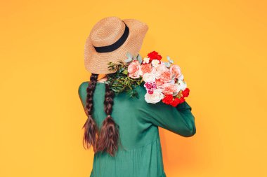 Yeşil elbiseli, kafasında hasır şapkası olan genç bir kadın omzuna sarı arka planda farklı çiçeklerin güzel bir karışımını koymuş..