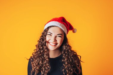Saçları kıvırcık ve kafasında Noel Baba şapkası olan güzel bir kadın. Noel havası. Sarı arkaplanda metin için boşluk