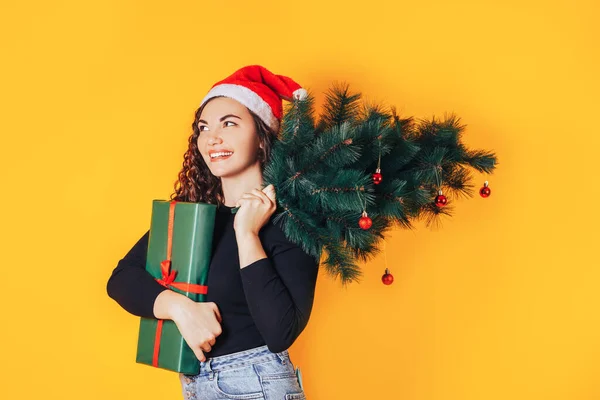 Sarı arka planda genç ve güzel bir kadın, kafasında Noel Baba şapkası var. Yeşil ambalajlı, kırmızı kurdeleli ve omzunda Noel ağacı olan bir kadın. Noel havası. Metin için yer — Stok fotoğraf