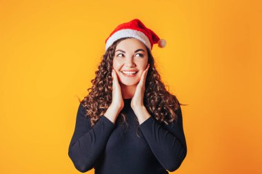 Kıvırcık saçlı ve başında Noel Baba şapkası olan güzel bir kadın. Kadın gülümsedi ve elleriyle yüzüne dokundu. Noel havası. Sarı arkaplanda metin için boşluk.