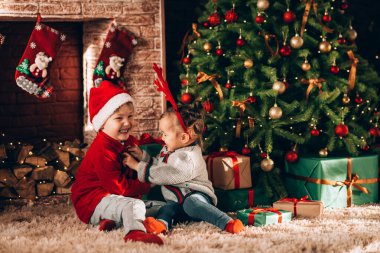 Erkek ve kız kardeşler yeşil paketler içinde hediyeler ve bir yayla oturuyorlar. Noel havası. Yeni yıl kostümlü çocuklar bir mucize bekliyorlar. Bir Noel ağacı ve şöminesinin arka planında.