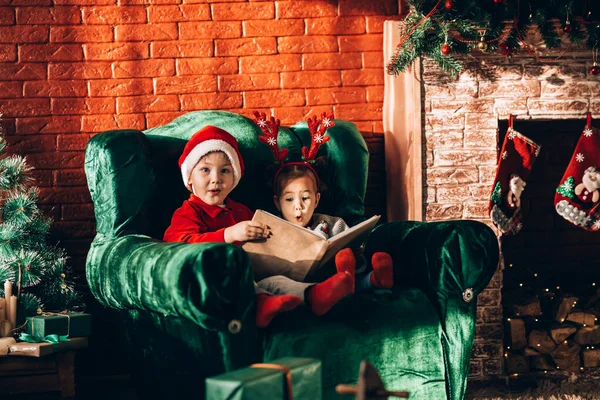 Brother and sister are sitting on a green armchair and reading a book. Christmas mood. Children in New Year\'s costumes waiting for a miracle.