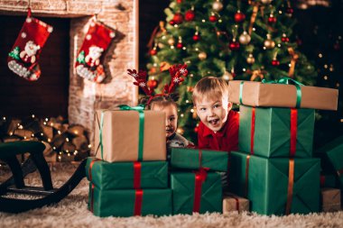 Erkek ve kız kardeşler yeşil paketler içinde hediyeler ve bir yayla oturuyorlar. Noel havası. Yeni yıl kostümlü çocuklar bir mucize bekliyorlar. Bir Noel ağacı ve şöminesinin arka planında.