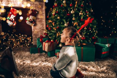 Süslü bir Noel ağacının önünde oturan küçük kız. Bir Noel ağacının arka planında, yeşil paketlenmiş kutular, fiyonk ve şömineyle. Noel havası.