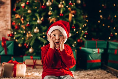 Çocuğun kafasında Noel Baba şapkası var. Arka planda yeşil ambalajlı ve fiyonklu hediyelerle süslenmiş büyük bir Noel ağacı var. Noel havası.