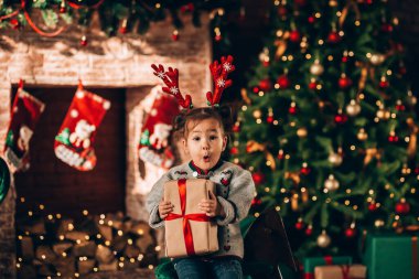 Süslü bir Noel ağacının önünde oturan küçük kız. Bir Noel ağacının arka planında, yeşil paketlenmiş kutular, fiyonk ve şömineyle. Noel havası.